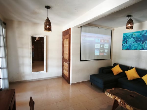 Sala con sofá azul, cojines amarillos, proyector encendido y mesa de madera rústica con luz natural