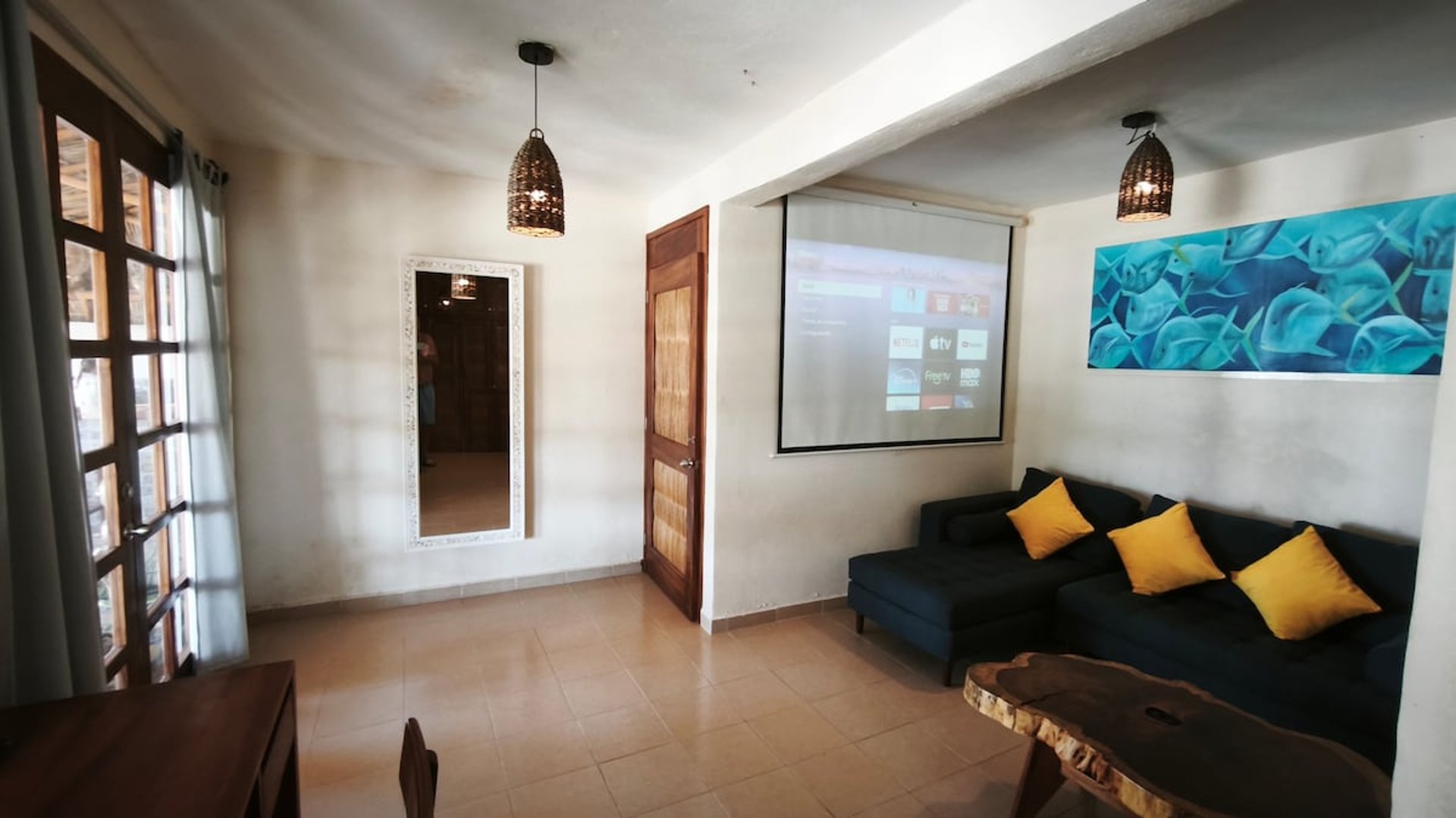 Sala con sofá azul, cojines amarillos, proyector encendido y mesa de madera rústica con luz natural