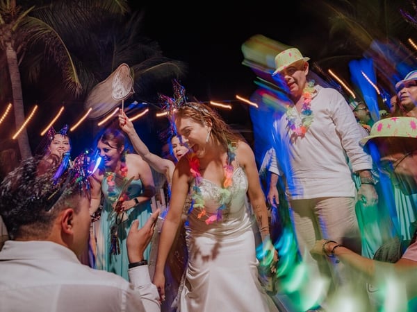 Novia bailando animada entre invitados con luces de neón y palmeras de fondo durante recepción nocturna de boda