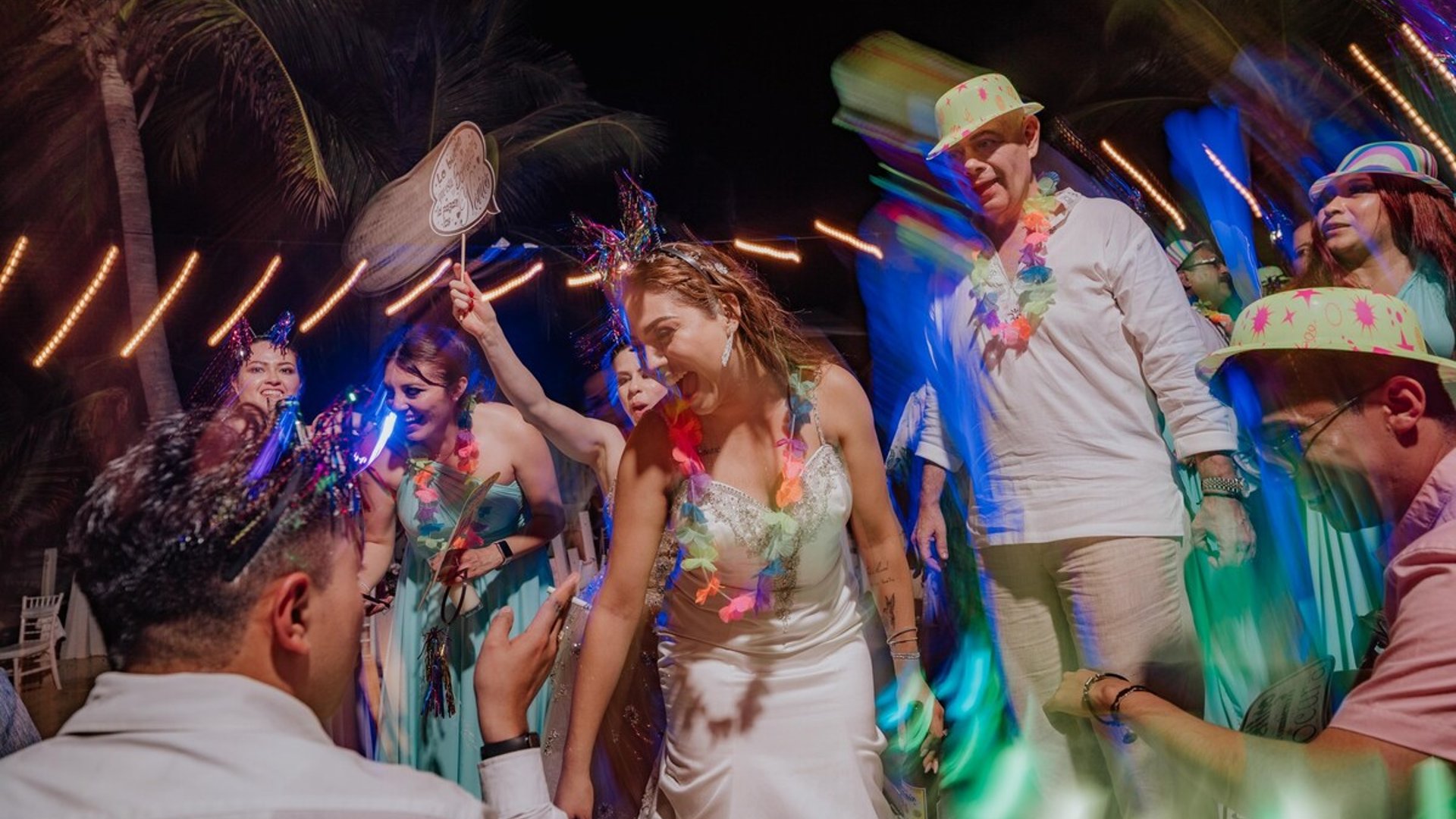 Novia bailando animada entre invitados con luces de neón y palmeras de fondo durante recepción nocturna de boda