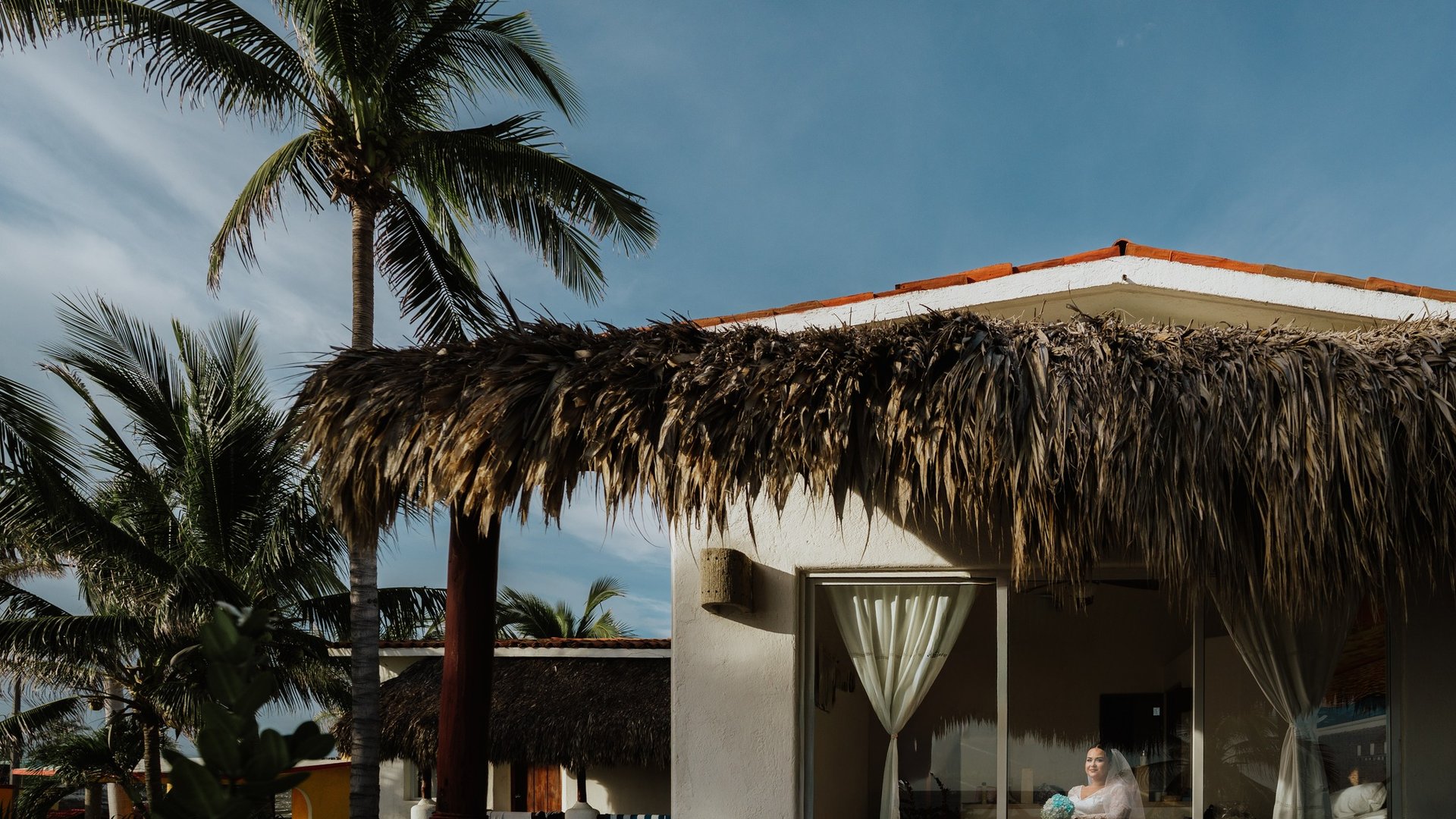 Villa frente al mar con palapa de palma y palmeras bajo cielo azul en ambiente tropical playero