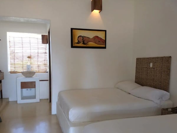 Habitación doble con camas individuales, cabeceras de mimbre, paredes blancas y pintura tropical en ambiente cálido