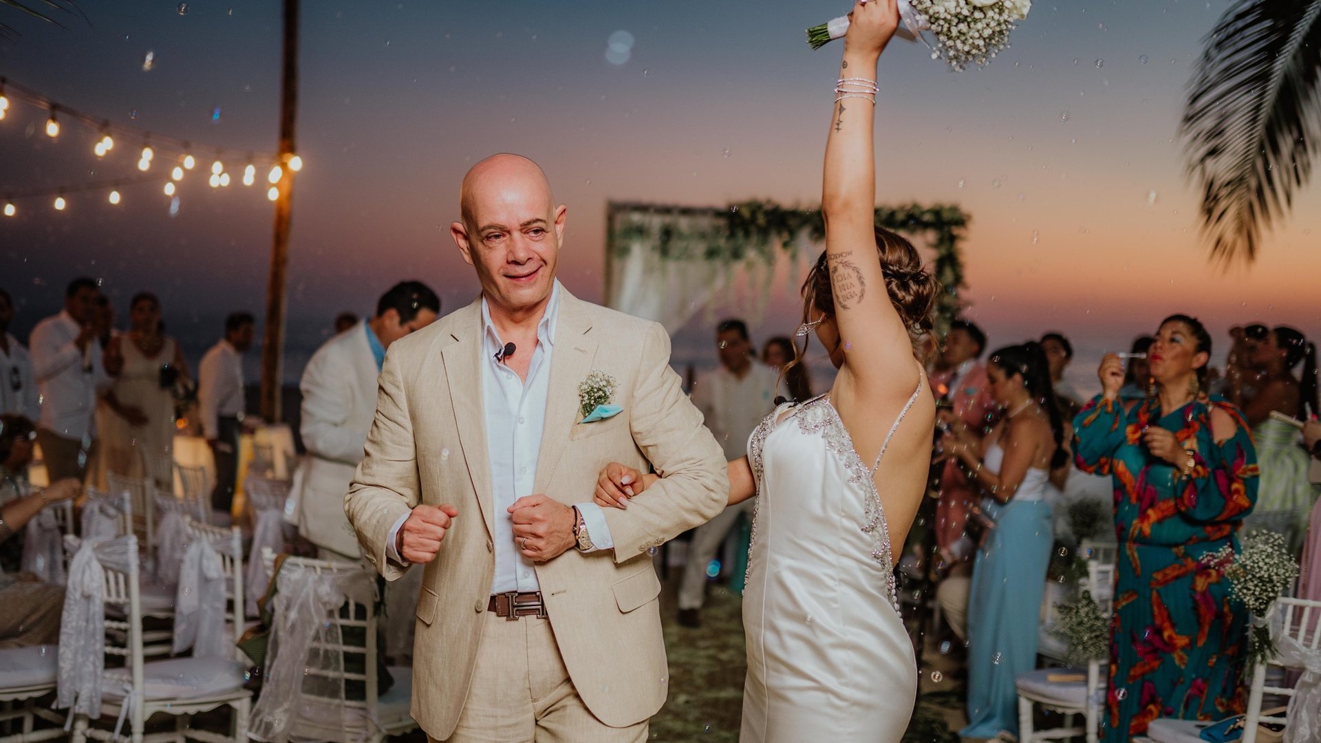 Celebración de boda al atardecer frente al mar con invitados y luces colgantes bajo palmeras