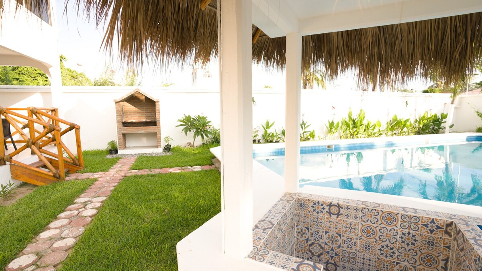 Jardín con pasto verde, asador de ladrillo, jacuzzi con mosaicos talavera y alberca bajo palapa en área exterior