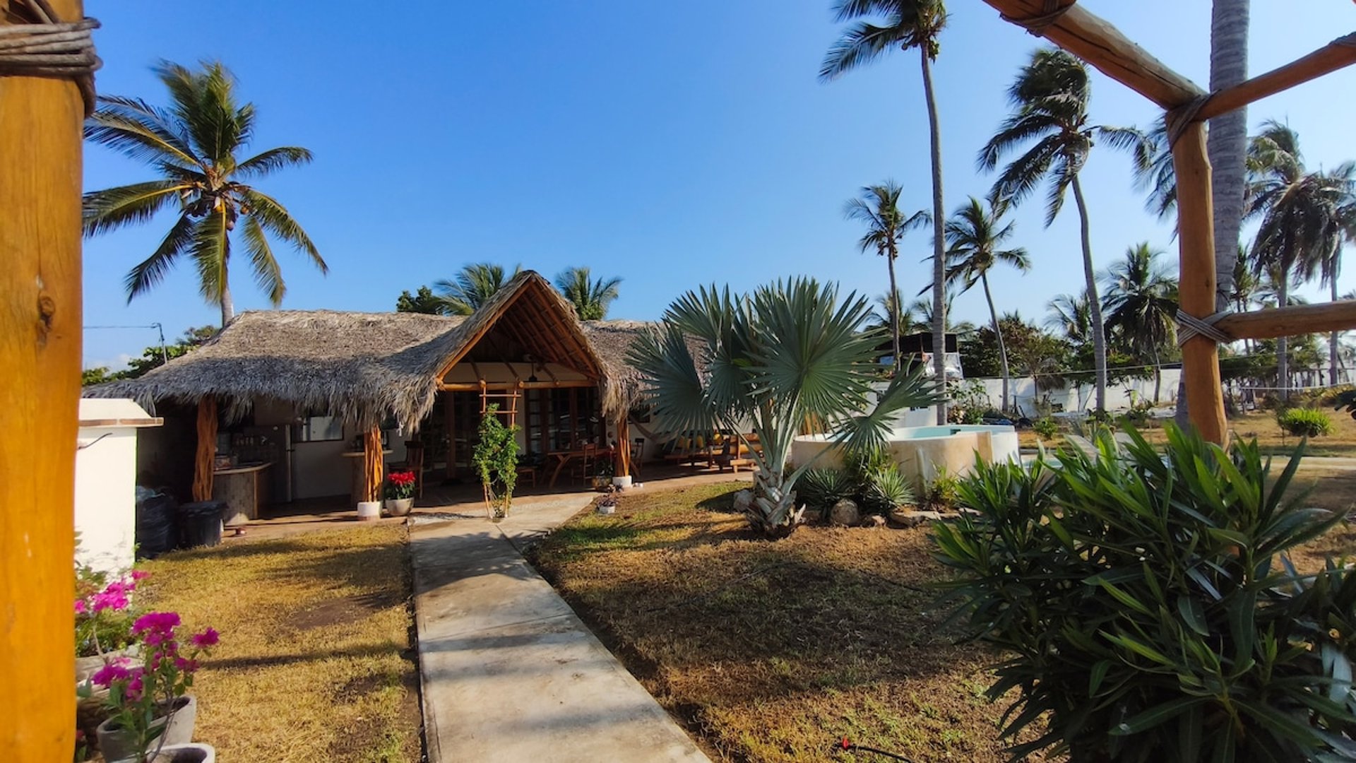 Vista general de palapa principal con jardín, palmeras y jacuzzi al fondo bajo cielo azul despejado