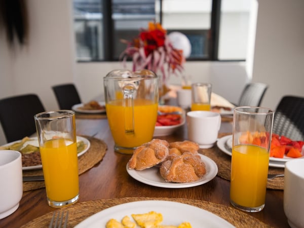 Mesa del comedor con desayuno servido: jugo de naranja, pan dulce, fruta y huevos en vajilla blanca