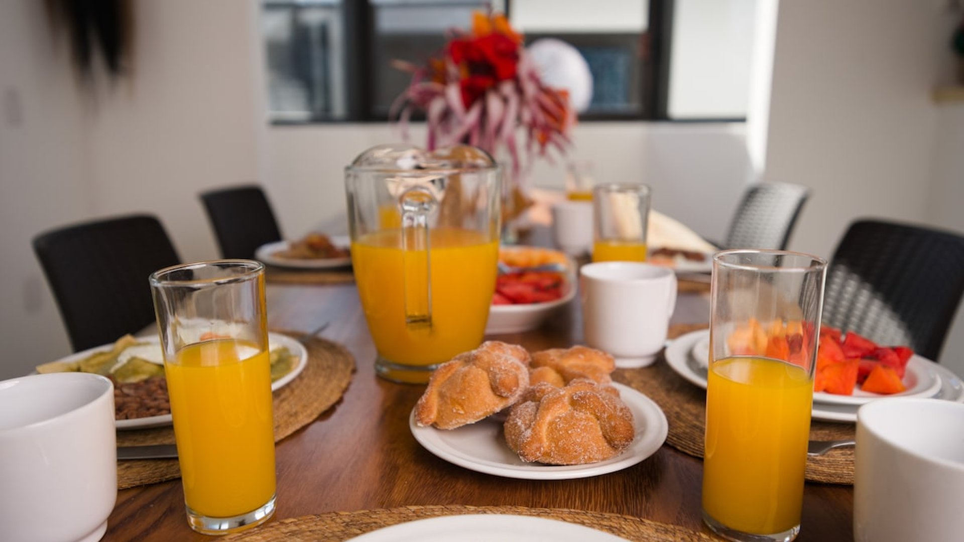 Mesa del comedor con desayuno servido: jugo de naranja, pan dulce, fruta y huevos en vajilla blanca