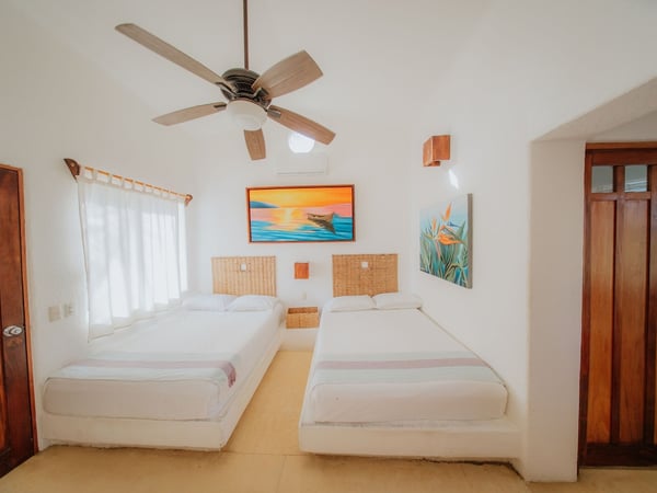 Recámara doble con dos camas matrimoniales, cabeceras de mimbre, pinturas tropicales y ventilador de techo en ambiente luminoso