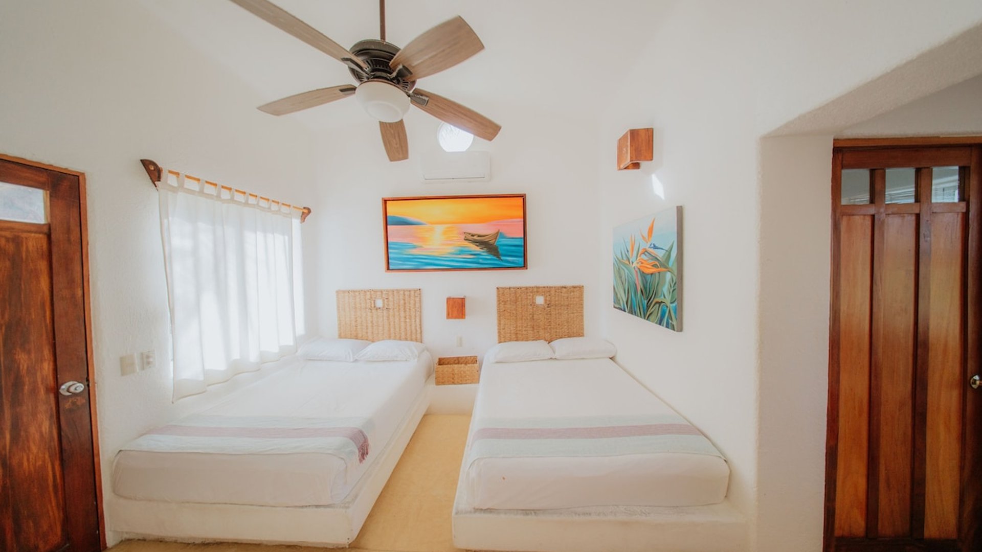 Recámara doble con dos camas matrimoniales, cabeceras de mimbre, pinturas tropicales y ventilador de techo en ambiente luminoso