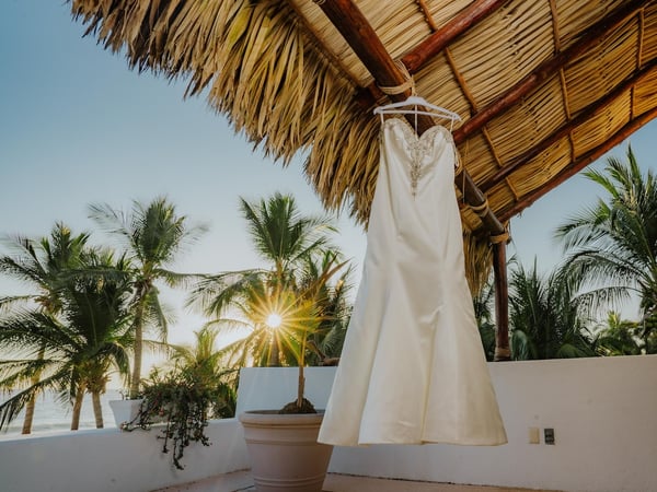 Vestido de novia colgado bajo palapa con vista al mar y palmeras iluminadas por el sol