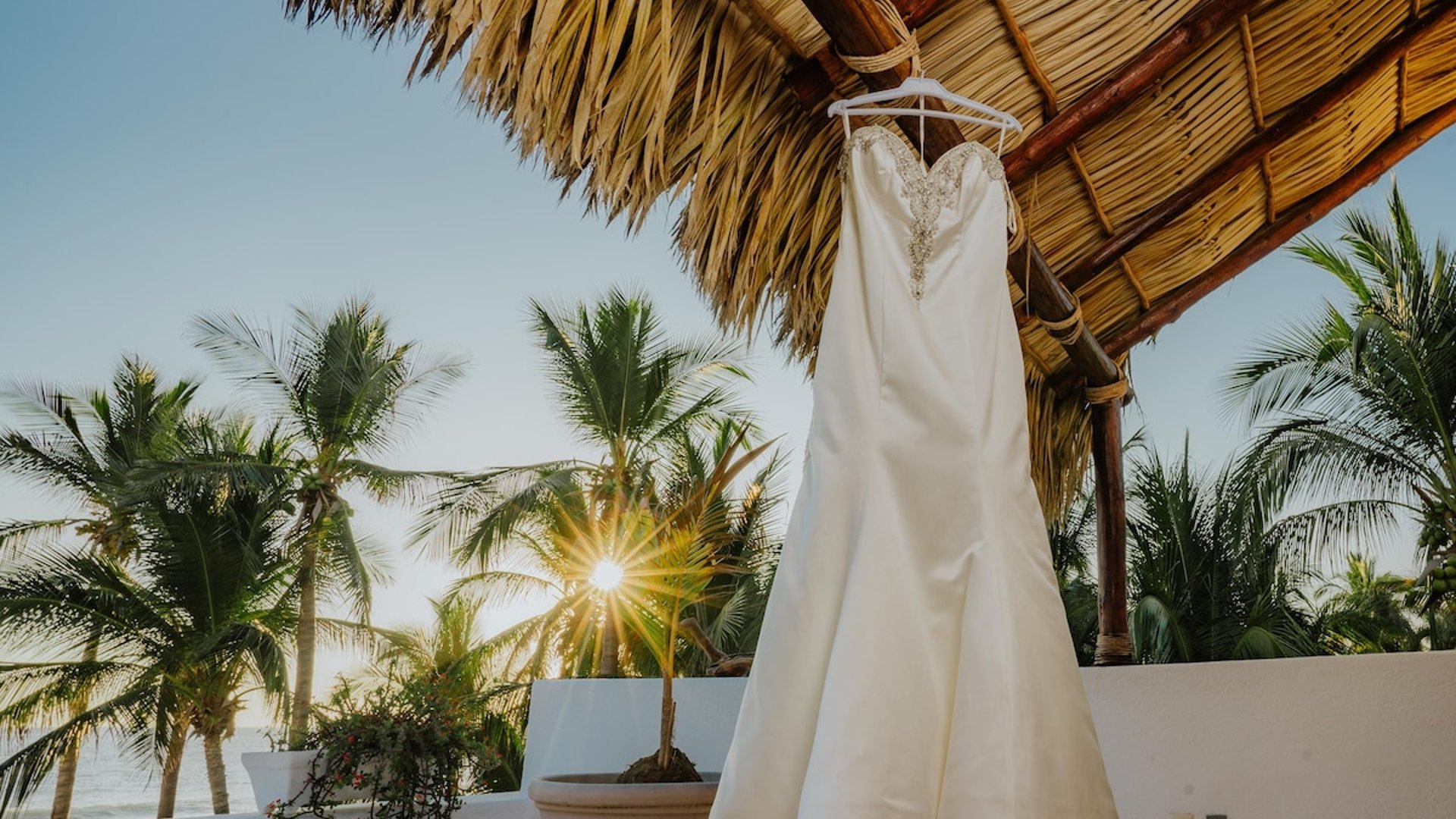 Vestido de novia colgado bajo palapa con vista al mar y palmeras iluminadas por el sol