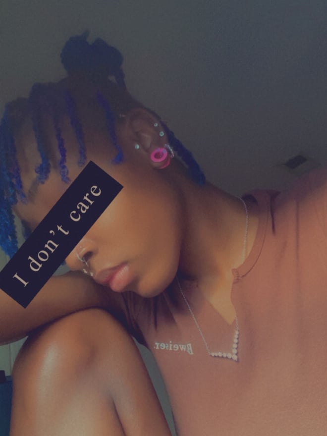 A woman with colorful braided hair sitting on the floor in a dimly lit room, wearing a casual top with text overlay.