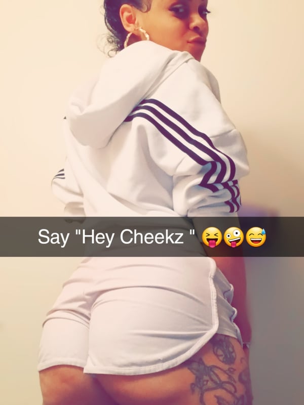 A woman in a white Adidas tracksuit stands smiling at the camera against a plain background, with tattoos visible on her arm.