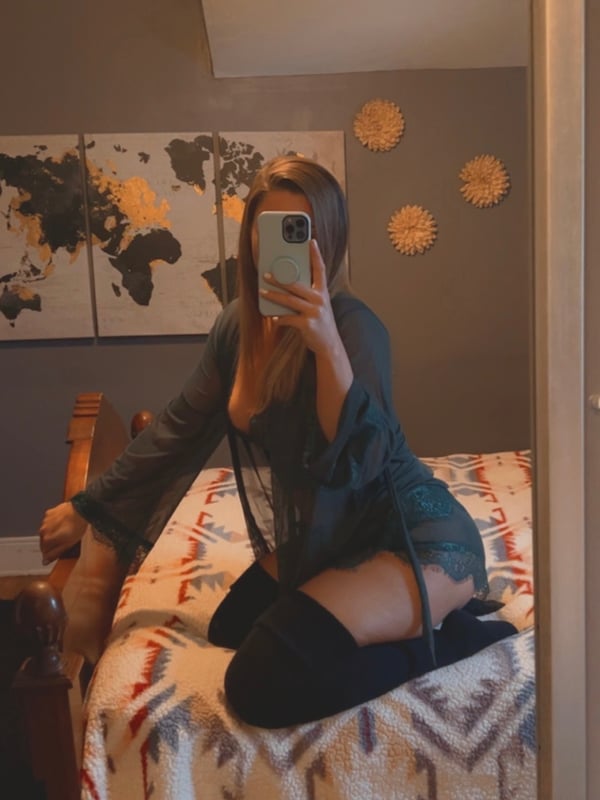 A woman taking a selfie in a gray top and thigh-high stockings while sitting on a bed in a bedroom with a world map poster.