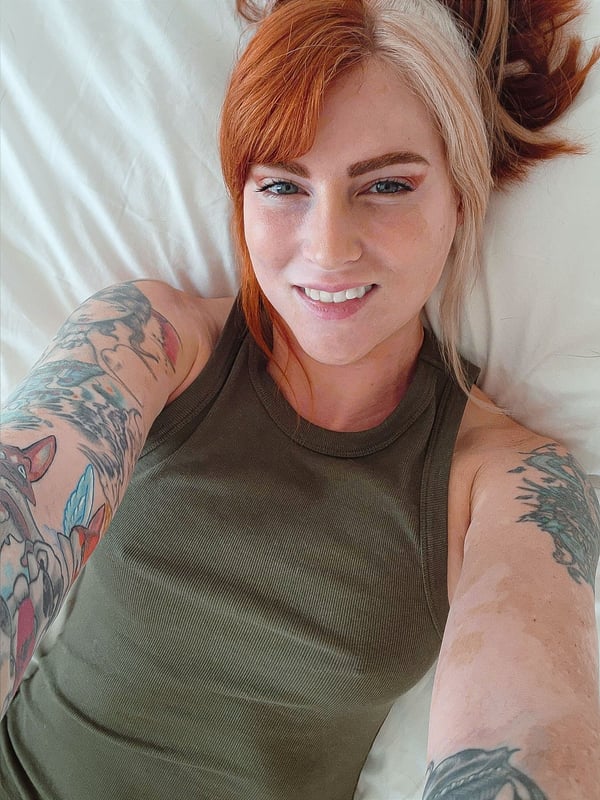 A smiling woman with dyed split-color hair and tattoos lies on a bed wearing a green tank top.