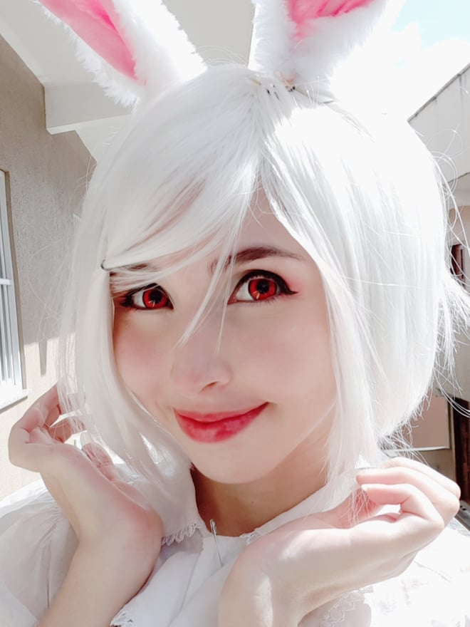 A person with a white wig, bunny ears, and red contact lenses smiling cutely outdoors on a balcony.