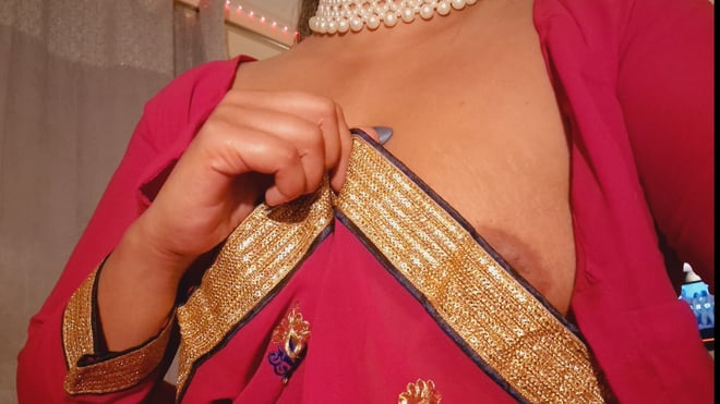 Woman in a red embroidered saree and pearl necklace posing indoors with decorative elements in the background.