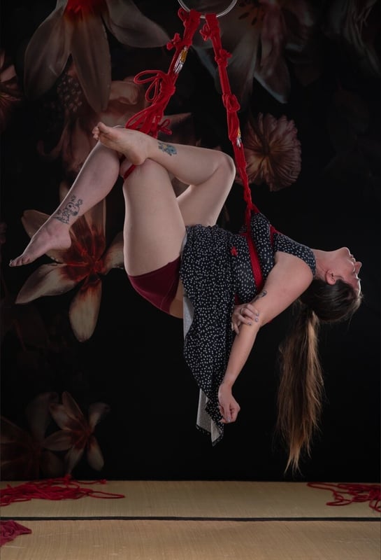 Woman suspended in red aerial silks amidst large flowers, wearing a short dress, posing dynamically in a studio setting.