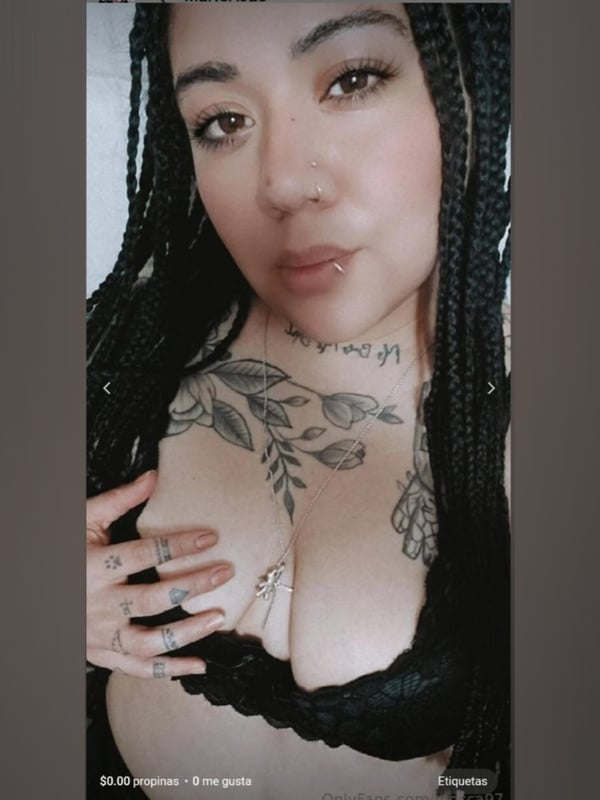 Close-up portrait of a woman with braided hair, tattoos on her chest, and a nose piercing, smiling softly.
