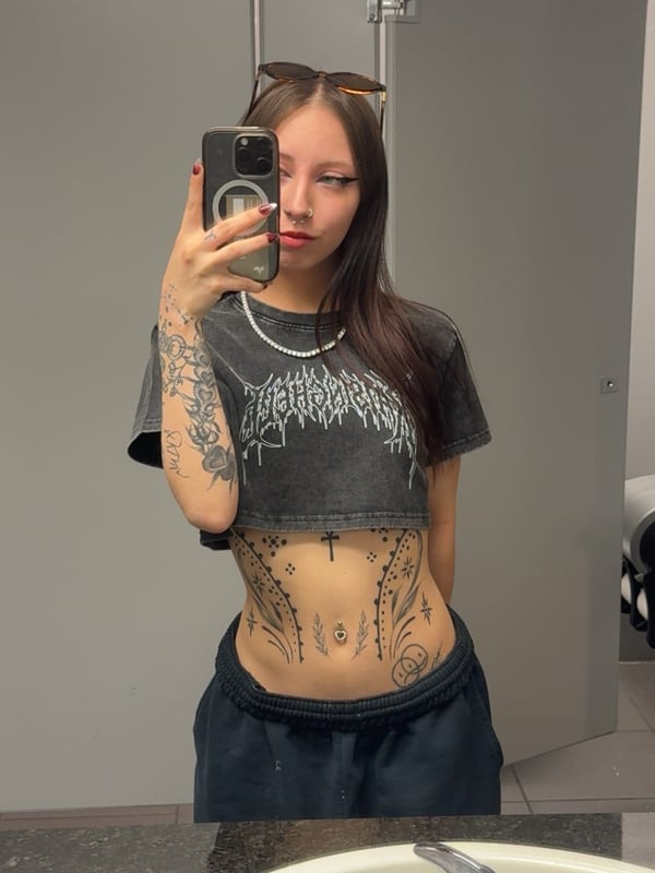 A close-up selfie of a woman with long brown hair, nose ring, and visible tattoos on her chest and neck, sitting inside a car.