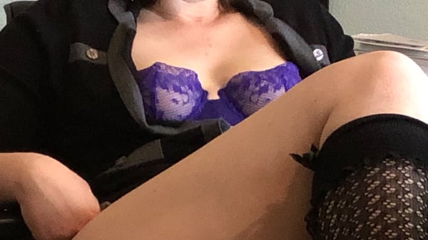 Woman in a black blazer over purple lace lingerie, sitting with legs crossed in an office-like setting.