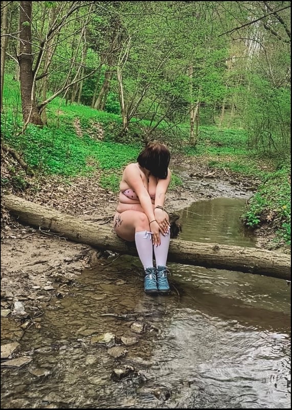 A woman sitting on a log by a stream in a lush green forest, wearing light-colored attire and boots.