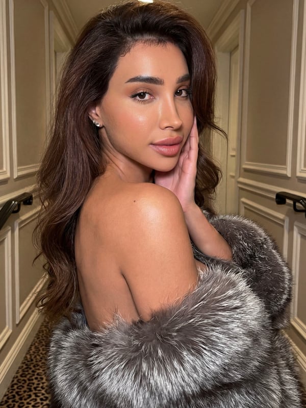 A woman with long brown hair and elegant makeup standing in a luxurious hallway, wearing a fur coat and touching her face.