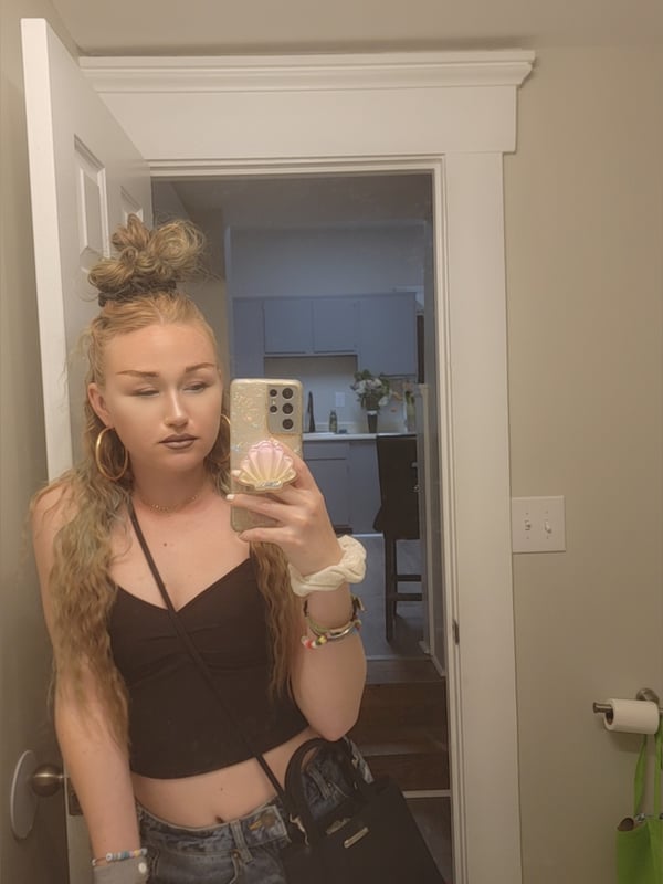 Young woman with blonde hair in a messy bun taking a mirror selfie in a casual top and jeans, standing in a doorway with a kitchen in the background.