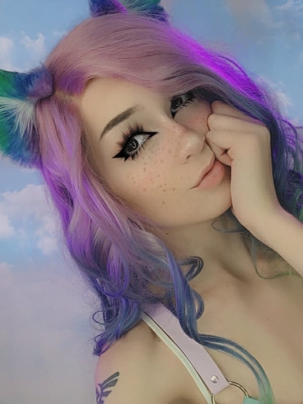 Young woman with colorful dyed hair, cat ears, and freckles posing playfully against a cloudy sky background.