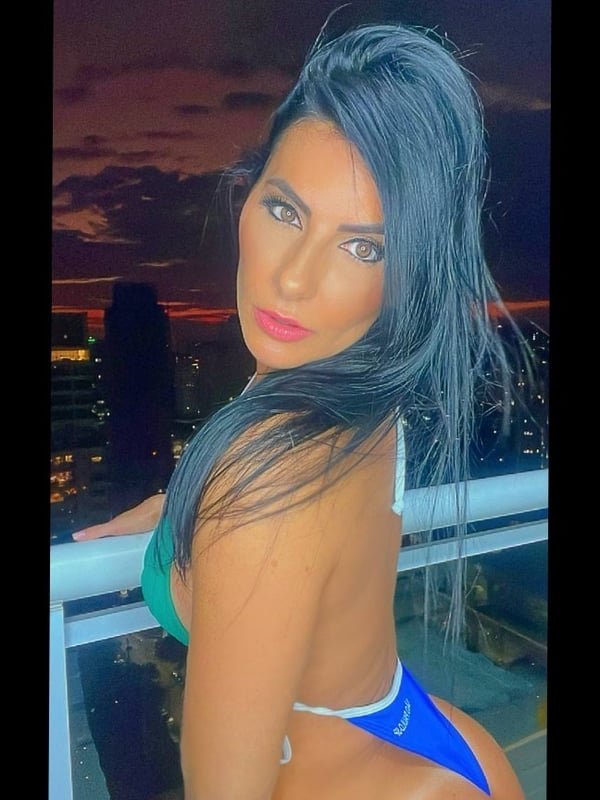 A woman with dark hair standing on a balcony at sunset, wearing a green and blue swimsuit, posing with a view of the city skyline behind her.