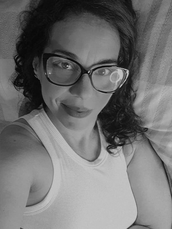 Close-up portrait of a woman with curly hair and glasses smiling on a bed, in black and white.