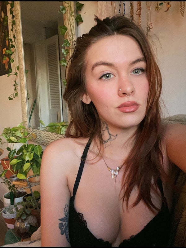 A young woman with brown hair smiling at the camera in a black top, sitting in a room with plants and bohemian decor.