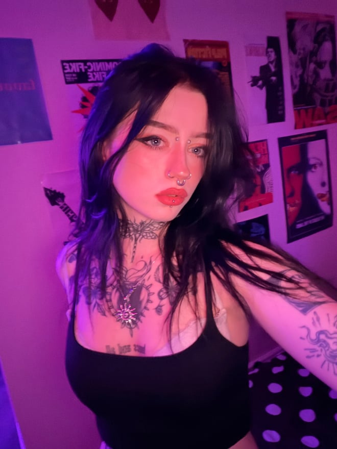 A tattooed woman with dark hair and facial piercings poses indoors under purple lighting, wearing a black top.