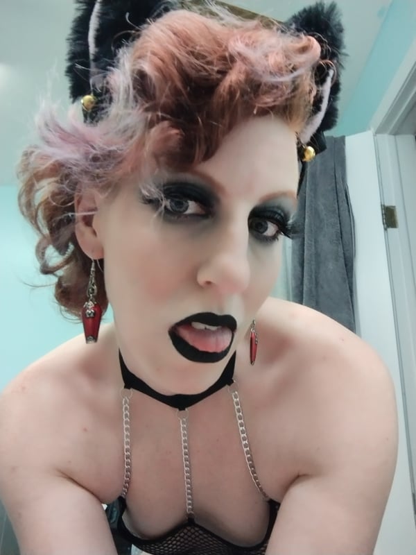 Woman with dyed wavy hair, cat ears headband, dramatic makeup, and choker necklace posing playfully in a bathroom mirror selfie.