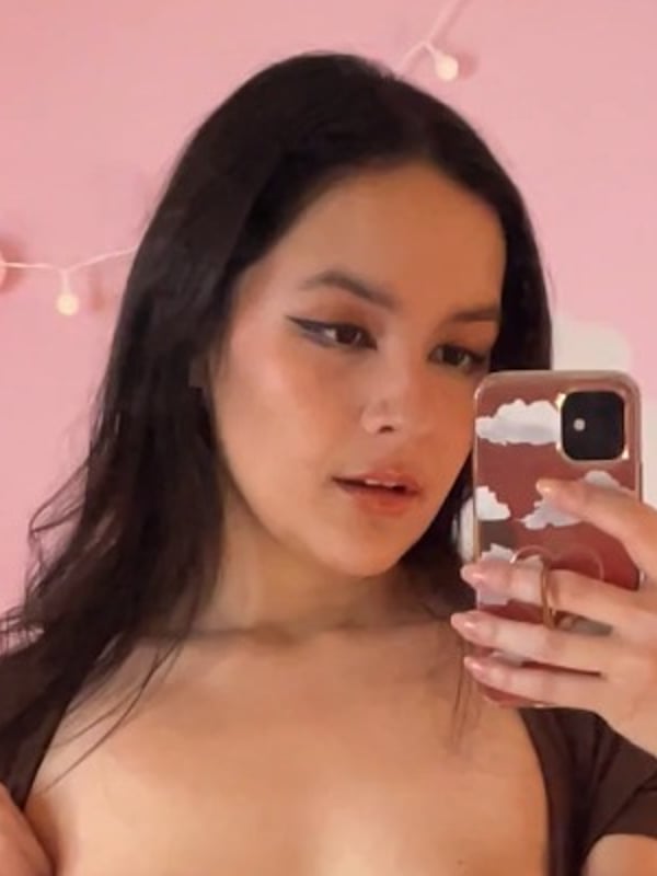A young woman taking a selfie in a pink room with string lights and clouds, holding her phone and smiling softly.