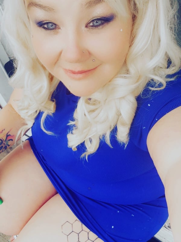 A close-up selfie of a woman with blonde wavy hair, blue eyes, and tattoos, smiling while wearing a blue top.