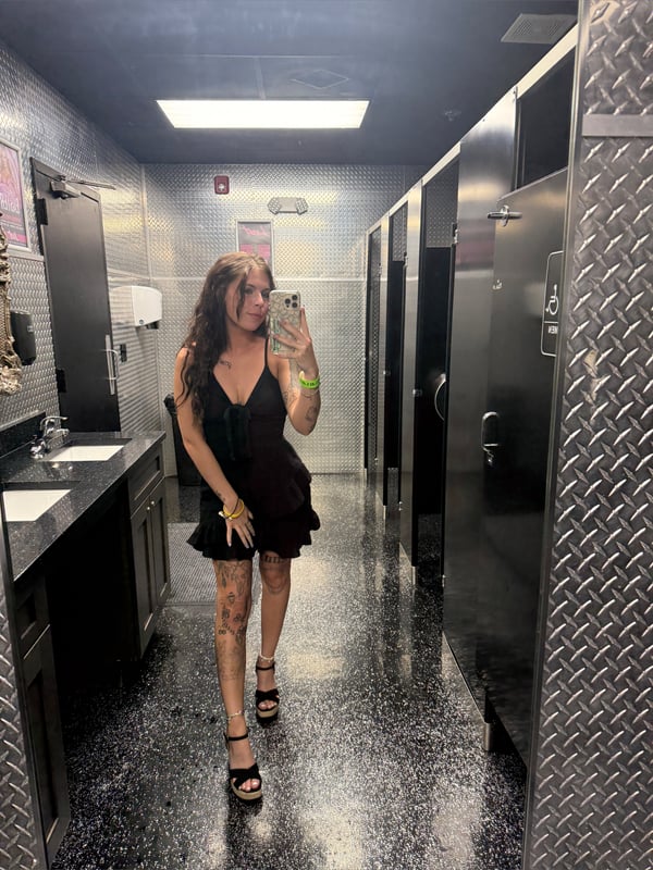 A woman taking a mirror selfie in a bathroom, wearing a short black dress and high heels.