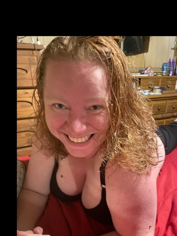 Woman with strawberry blonde wavy hair smiling happily while reclining on a red bed in a black top, with wooden furniture in the background.
