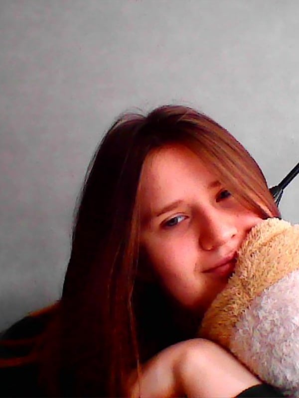 Young woman with long reddish-brown hair smiling while cuddling a large teddy bear against a plain background.