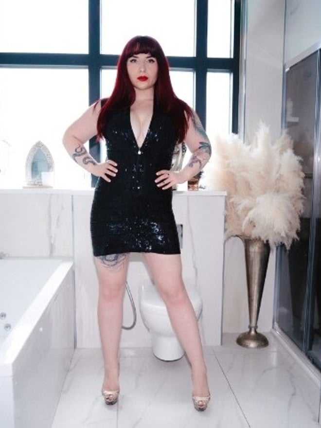 A woman with red hair standing confidently in a sparkling black dress in a modern bathroom setting with a bathtub.