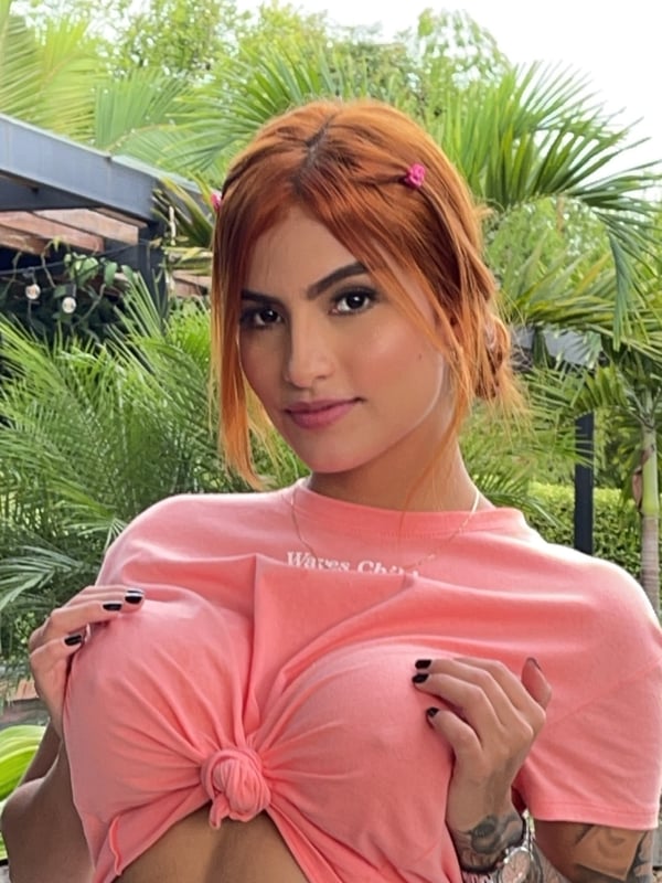 A person with red hair wearing a pink top, posing outdoors with green plants in the background.