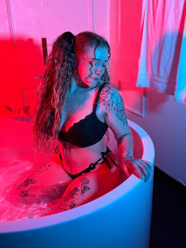 Woman with wavy hair in pigtails, sitting in a bathtub with pink and blue lighting, wearing a black bikini top.