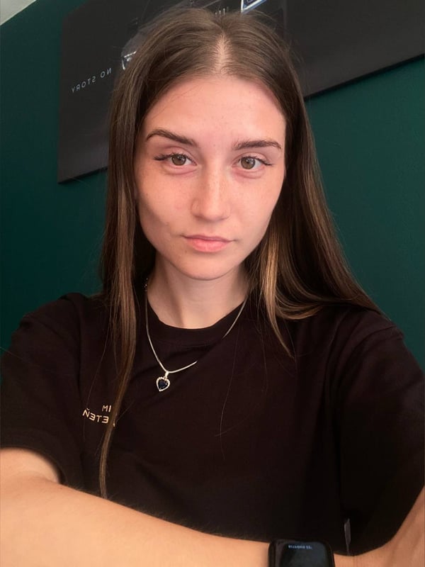Young woman with long brown hair taking a selfie in a dark top against a green wall.