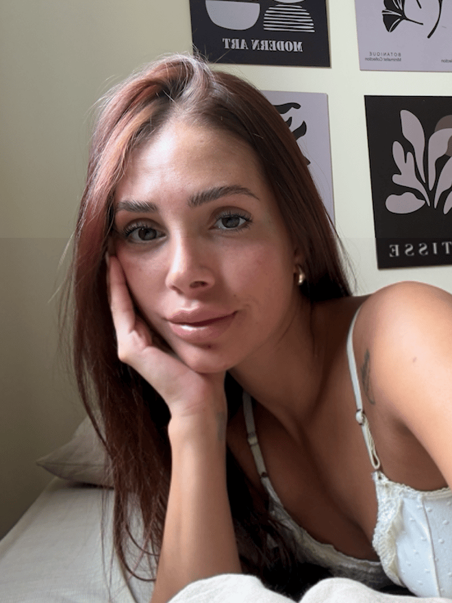 A smiling woman with long reddish-brown hair taking a selfie from above while lying on white bedding, wearing a white lace-trimmed top.