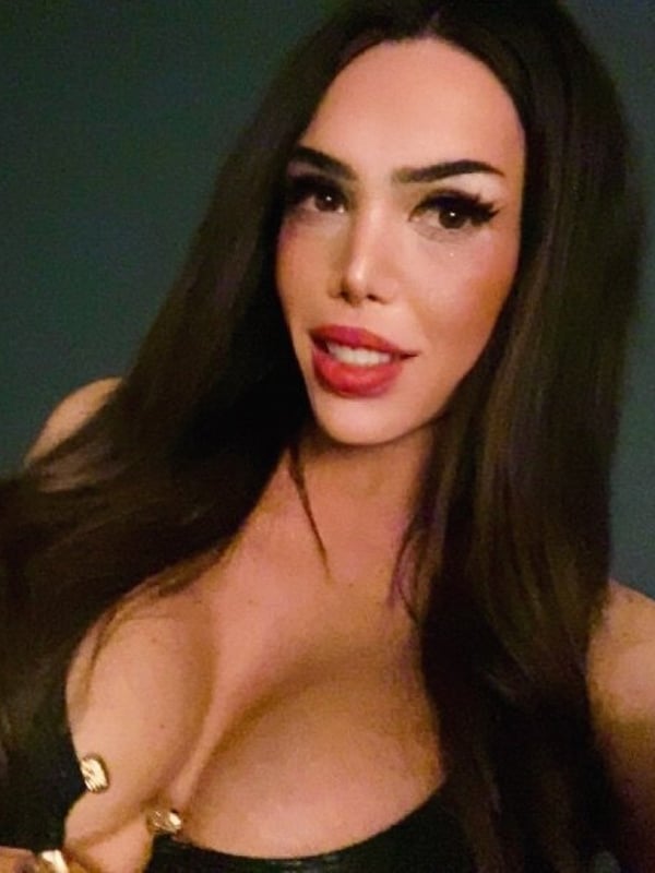 A woman with long dark hair and bold makeup posing confidently in a low-cut black top, showing cleavage.