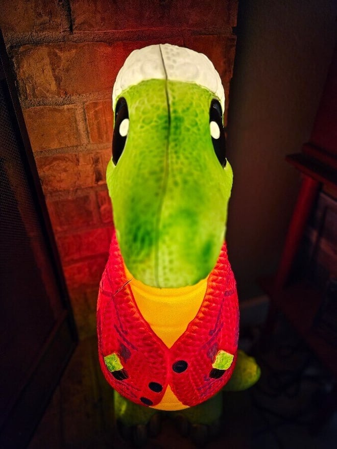 Green plush dinosaur toy dressed in a colorful sweater vest, standing against a brick wall.