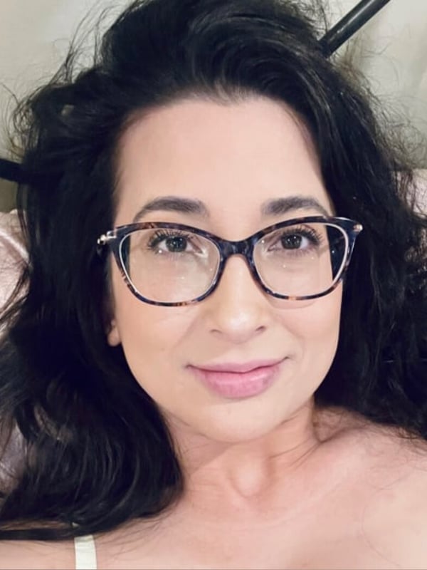 A woman with dark hair and glasses smiling softly while lying on a bed, dressed in a light top against a neutral background.
