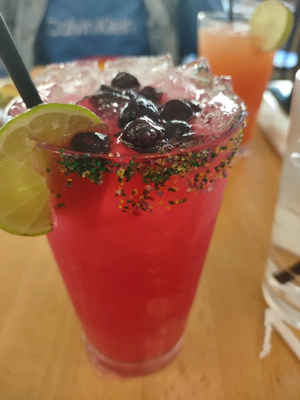Close-up of a vibrant pink margarita garnished with blueberries, lime slices, and green sugar rim on a restaurant table next to another drink.