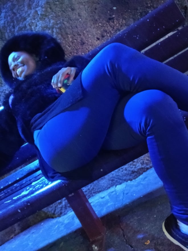 A woman lounging on a wooden bench outdoors in a fur-trimmed jacket, blue pants, and sneakers against a rocky blue-toned background.