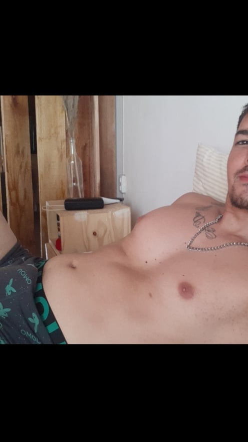 A man lounging shirtless on a bed in a room with wooden furniture and a plant.